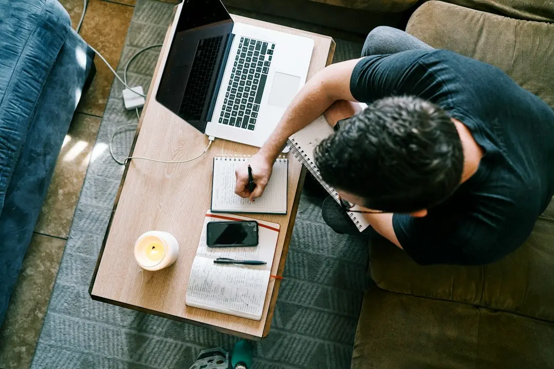 A person takes notes in a notebook at a coffee table, next to a laptop and a phone, while comparing IPTV providers before an IPTV Free Trial